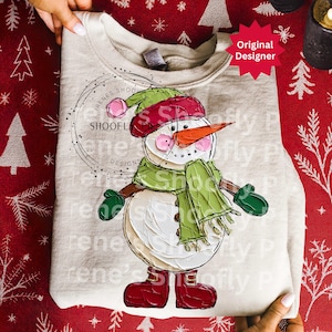 Op de afbeelding: Een beige sweatshirt met een geschilderd sneeuwmanontwerp. De sneeuwman draagt een groene sjaal, muts en wanten, met een wortelneus en rode laarzen. Het ontwerp bevat de tekst "RENES SHOOFL DESIGNS" en "Shoofly P".