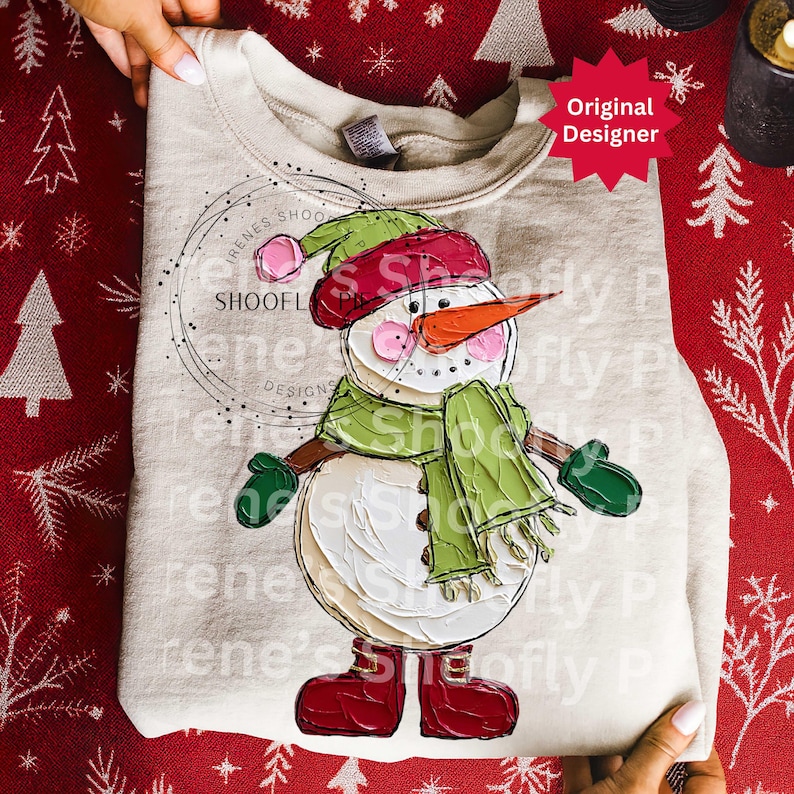 Op de afbeelding: Een beige sweatshirt met een geschilderd sneeuwmanontwerp. De sneeuwman draagt een groene sjaal, muts en wanten, met een wortelneus en rode laarzen. Het ontwerp bevat de tekst "RENES SHOOFL DESIGNS" en "Shoofly P".