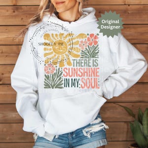 May include: White hoodie with a graphic design featuring a smiling sun, flowers, and the text "THERE IS SUNSHINE IN MY SOUL." The design includes yellow, pink, green, and coral colors. The hoodie has a front pocket and a drawstring hood.