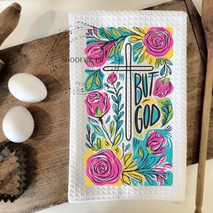 May include: White kitchen towel with a colorful floral design and the text "But God" in a black cross shape.