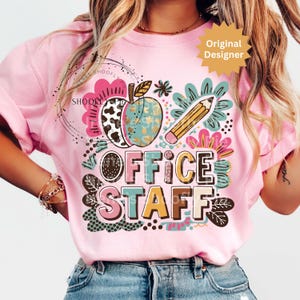 Può includere: T-shirt rosa chiaro con un design grafico colorato raffigurante una mela, una matita e le parole "OFFICE STAFF". Il design include elementi floreali e fantasia in vari colori, creando un'estetica giocosa e allegra.