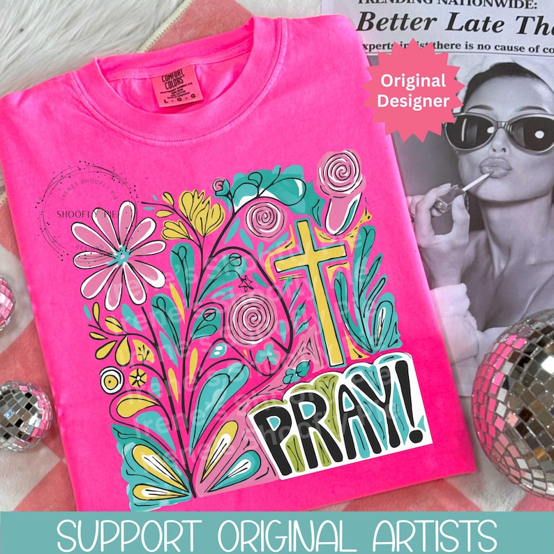 May include: A hot pink t-shirt featuring a colorful floral design with a yellow cross and the word "PRAY!" in bold letters. The design includes flowers, leaves, and swirls in various colors. The shirt also has the text "SUPPORT ORIGINAL ARTISTS".