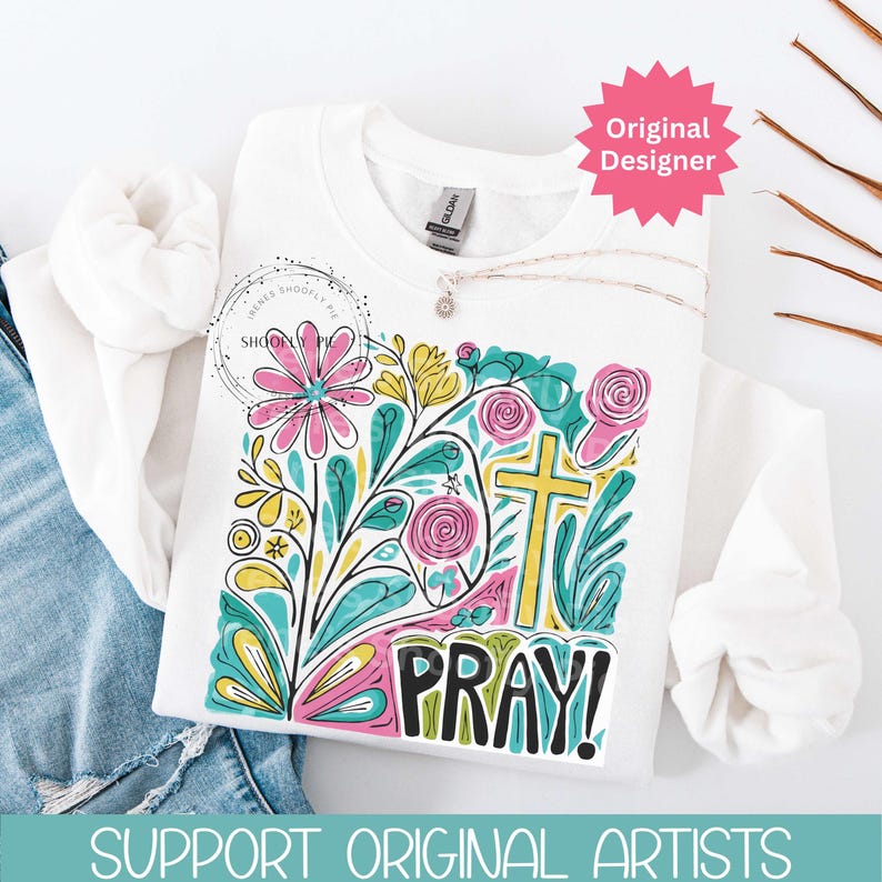 May include: White sweatshirt with a colorful floral design, a yellow cross, and the word "PRAY!" in black. The design includes pink, teal, and yellow flowers and leaves. A silver necklace with a pendant is draped across the sweatshirt.