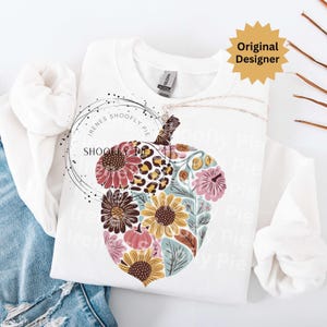May include: White sweatshirt featuring a floral acorn design. The acorn is filled with sunflowers, pink flowers, and a leopard print pattern. The text "SHOOFY PIE" is in a circle on the left side of the sweatshirt. The words "Original Designer" are in a yellow starburst.