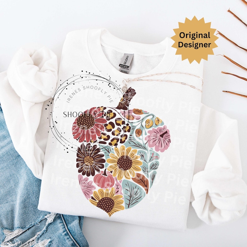 May include: White sweatshirt featuring a floral acorn design. The acorn is filled with sunflowers, pink flowers, and a leopard print pattern. The text "SHOOFY PIE" is in a circle on the left side of the sweatshirt. The words "Original Designer" are in a yellow starburst.