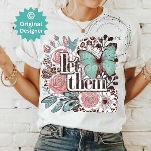 May include: White t-shirt with a floral and butterfly design. The text "Let them" is printed on the shirt.