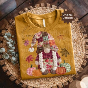 Peut inclure: T-shirt jaune moutarde avec un motif de gnome. Le gnome porte un chapeau en patchwork, des tresses et une robe &agrave; motifs floraux. Des citrouilles et des feuilles d'automne sont &eacute;galement repr&eacute;sent&eacute;es. Le texte "SHOOFY PIE" est visible.