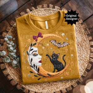 Op de afbeelding: Mosterdgeel T-shirt met een geborduurd ontwerp met een halve maan, een zwarte kat en een vleermuis. De maan heeft een gezicht en is omringd door sterren. Bovenop zit een bordeauxrode strik. De tekst "SHOOFly PIE DESIGN" staat op de maan.
