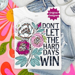 Op de afbeelding: Wit T-shirt met een bloemenontwerp en de tekst "DON'T LET THE HARD DAYS WIN". De bloemen hebben luipaardprintcentra en zijn in tinten roze, paars en wit. Het shirt wordt getoond op een blauwe spijkerbroek.