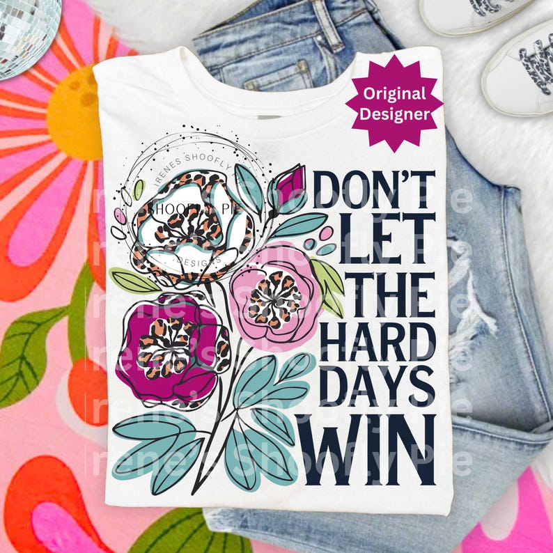 Op de afbeelding: Wit T-shirt met een bloemenontwerp en de tekst "DON'T LET THE HARD DAYS WIN". De bloemen hebben luipaardprintcentra en zijn in tinten roze, paars en wit. Het shirt wordt getoond op een blauwe spijkerbroek.