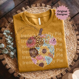 May include: Mustard yellow t-shirt featuring a floral pumpkin design. The pumpkin is composed of sunflowers, other flowers, and a leopard print section. The text "Irene's Shoofly Pie" is printed on the shirt. A pink starburst with the words "Original Designer" is in the upper right corner.