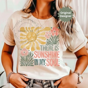 May include: Cream-colored t-shirt featuring a colorful graphic design. The design includes a smiling sun, flowers, and the text "THERE IS SUNSHINE IN MY SOUL." The shirt has short sleeves and a round neckline.