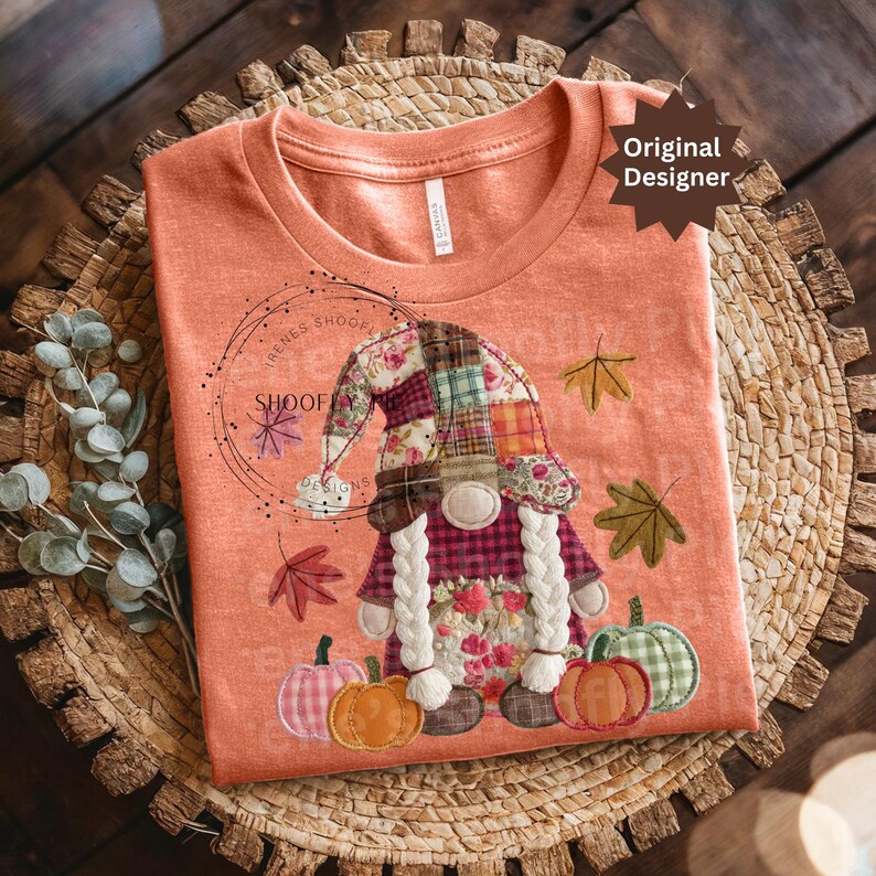 Peut inclure: T-shirt p&ecirc;che avec un motif de gnome fantaisiste. Le gnome porte un chapeau et une barbe en patchwork, avec des feuilles d'automne et des citrouilles. Le texte "SHOOFY DESIGNS" est &eacute;galement visible. Le t-shirt est pr&eacute;sent&eacute; sur une surface tiss&eacute;e.