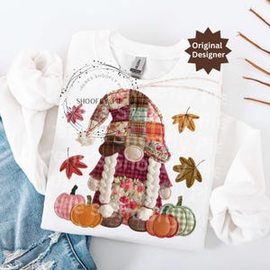 Peut inclure: Sweat-shirt blanc &agrave; manches longues avec un motif de gnome. Le gnome porte un chapeau en patchwork et des tresses, entour&eacute; de feuilles d'automne et de citrouilles. Le texte "SHOOFY PIE" est imprim&eacute; sur le sweat-shirt. Les mots "Original Designer" sont &eacute;galement imprim&eacute;s.