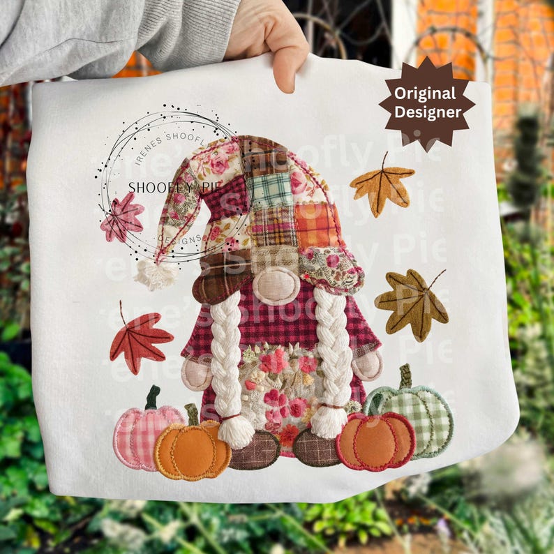 Peut inclure: Un sac fourre-tout blanc avec un motif de gnome d'automne. Le gnome porte un chapeau en patchwork et une chemise &agrave; carreaux, avec de longues tresses blanches. Des citrouilles et des feuilles d'automne entourent le gnome. Le texte "Original Designer" est en haut &agrave; droite.