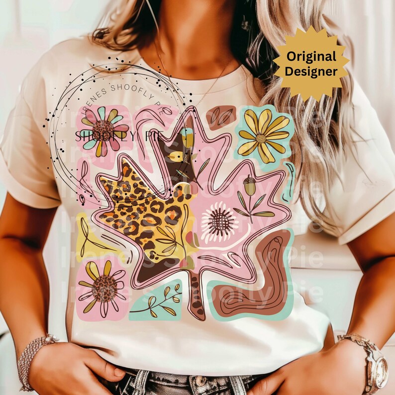 May include: A cream-colored t-shirt featuring a colorful leaf design with floral and leopard print accents. The design includes pink, yellow, brown, and teal elements. The words "Original Designer" are printed on a yellow starburst.