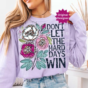 Op de afbeelding: Een lichtpaars shirt met lange mouwen met een bloemenontwerp en de tekst "DON'T LET THE HARD DAYS WIN". Het bloemenontwerp bevat witte, roze en paarse bloemen met luipaardprint accenten.