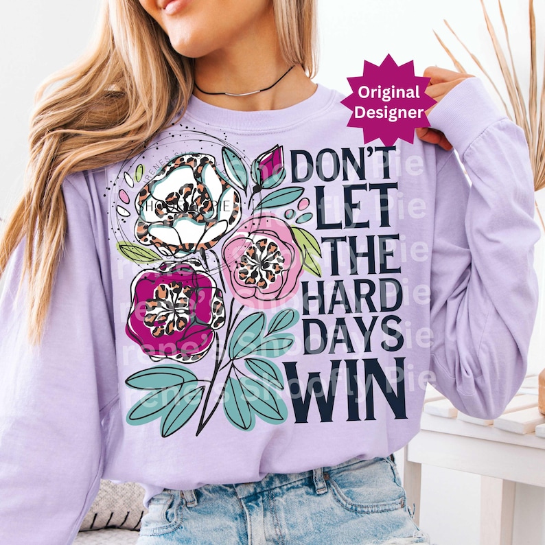 Op de afbeelding: Een lichtpaars shirt met lange mouwen met een bloemenontwerp en de tekst "DON'T LET THE HARD DAYS WIN". Het bloemenontwerp bevat witte, roze en paarse bloemen met luipaardprint accenten.
