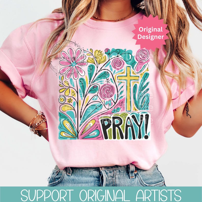 May include: Pink t-shirt with a colorful floral and cross design. The graphic includes the word "PRAY!" in bold letters. A pink starburst shape with the words "Original Designer" is in the upper right corner.
