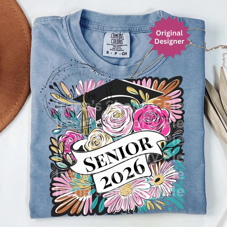 以下が含まれることがあります： 卒業式の花柄デザインが施された、ライトブルーのComfort Colors Tシャツ。 デザインには卒業キャップ、「SENIOR 2026」の文字、ピンクと白のバラが含まれています。 シャツには「Original Designer」と書かれたタグが付いています。