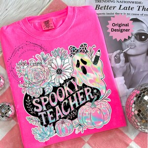 May include: A bright pink Comfort Colors sweatshirt featuring a floral and ghost design with the text "Spooky Teacher". The design includes pumpkins, flowers, and a ghost holding a pencil. The sweatshirt has a crew neck.