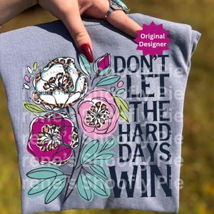 Op de afbeelding: Lichtblauw T-shirt met een bloemenontwerp en luipaardprint accenten. Het ontwerp bevat roze, witte en paarse bloemen met groene bladeren. De tekst "DON'T LET THE HARD DAYS WIN" is in donkerblauw gedrukt.