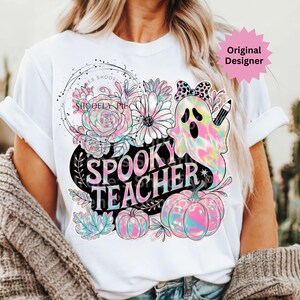 May include: White t-shirt featuring a colorful graphic design with the words "Spooky Teacher". The design includes a ghost, pumpkins, flowers, and the text "Shoofly Pie Designs". The design has a pastel rainbow color scheme.