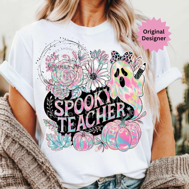 May include: White t-shirt featuring a colorful graphic design with the words "Spooky Teacher". The design includes a ghost, pumpkins, flowers, and the text "Shoofly Pie Designs". The design has a pastel rainbow color scheme.