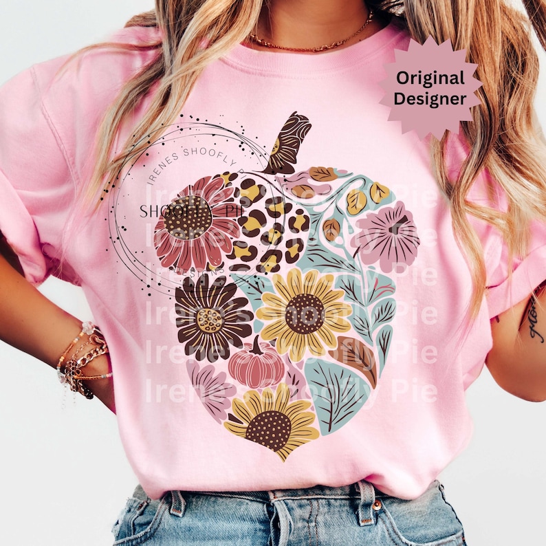 May include: Pink t-shirt featuring a floral pumpkin design. The pumpkin is filled with sunflowers, leaves, and a leopard print pattern. The text "Irenes Shoofly Pie" is printed on the shirt, along with the words "Original Designer".