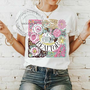 May include: White t-shirt featuring a floral design and the word "REALTOR" in a circle. The design includes a house, flowers, and leaves in shades of pink, green, and yellow.