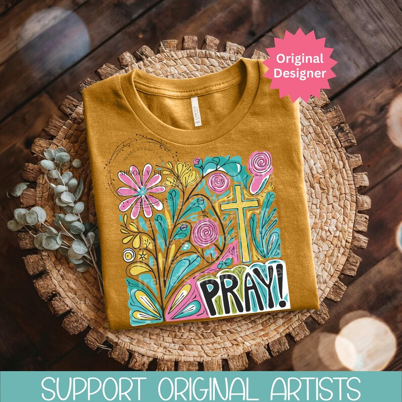 May include: Mustard-colored t-shirt with a floral and cross design. The graphic includes pink, blue, yellow, and green elements, with the word "PRAY!" in a bold font. The shirt is displayed on a woven mat.