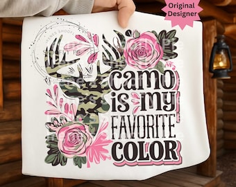 May include: White t-shirt with a camo deer head design, pink roses, and foliage. The text "Camo is my favourite colour" is in a bold, multi-coloured font. The design includes a circular logo and a pink star that says "Original Designer".