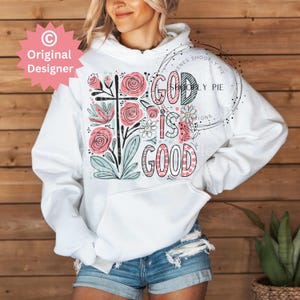 以下が含まれることがあります： 白いフード付きのスウェットシャツに、花柄のデザインと「God is Good」というピンクと黒の文字が書かれています。