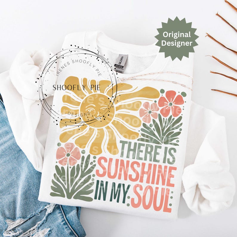 May include: White sweatshirt with a colorful graphic design. The design features a smiling sun, flowers, and the text "THERE IS SUNSHINE IN MY SOUL" in various colors. The sweatshirt is folded on a denim surface.