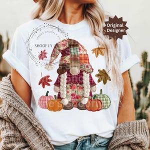 Peut inclure: T-shirt blanc avec un motif de gnome d'automne. Le gnome porte un chapeau patchwork, des tresses et des d&eacute;tails floraux. Des citrouilles et des feuilles d'automne entourent le gnome. Le texte "Original Designer" est visible.