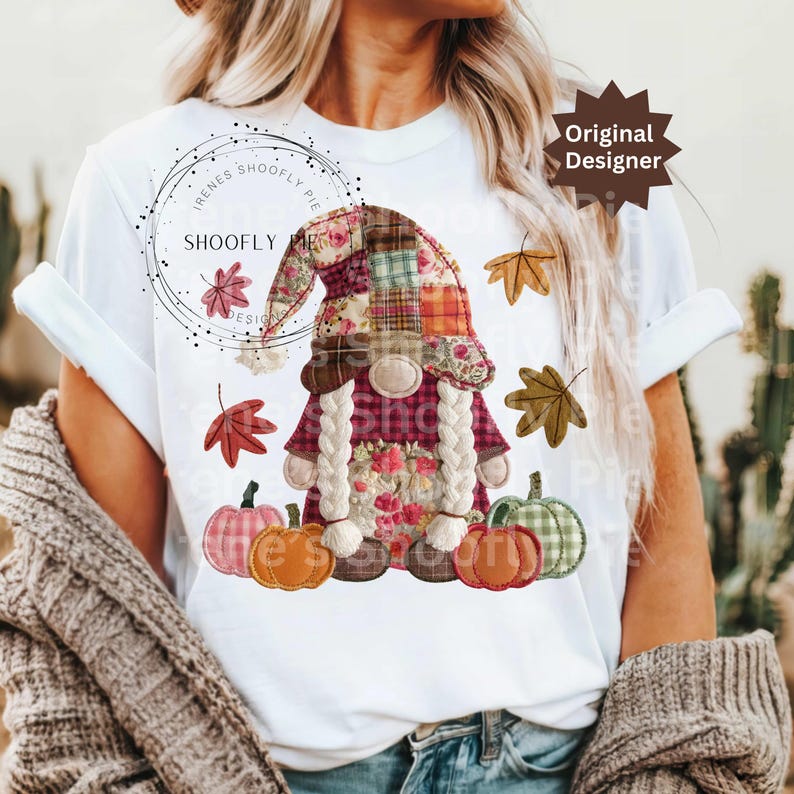 Peut inclure: T-shirt blanc avec un motif de gnome d'automne. Le gnome porte un chapeau patchwork, des tresses et des d&eacute;tails floraux. Des citrouilles et des feuilles d'automne entourent le gnome. Le texte "Original Designer" est visible.