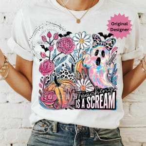May include: White t-shirt featuring a colorful graphic with pumpkins, flowers, and a ghost holding a pencil. The text "Teaching is a Scream" is displayed below the ghost. The design includes bats and a leopard print pattern.