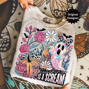 May include: A beige sweatshirt featuring a colorful graphic with a ghost, pumpkins, flowers, and the text "Teaching is a Scream." The design includes pink roses, daisies, and a leopard print bow.