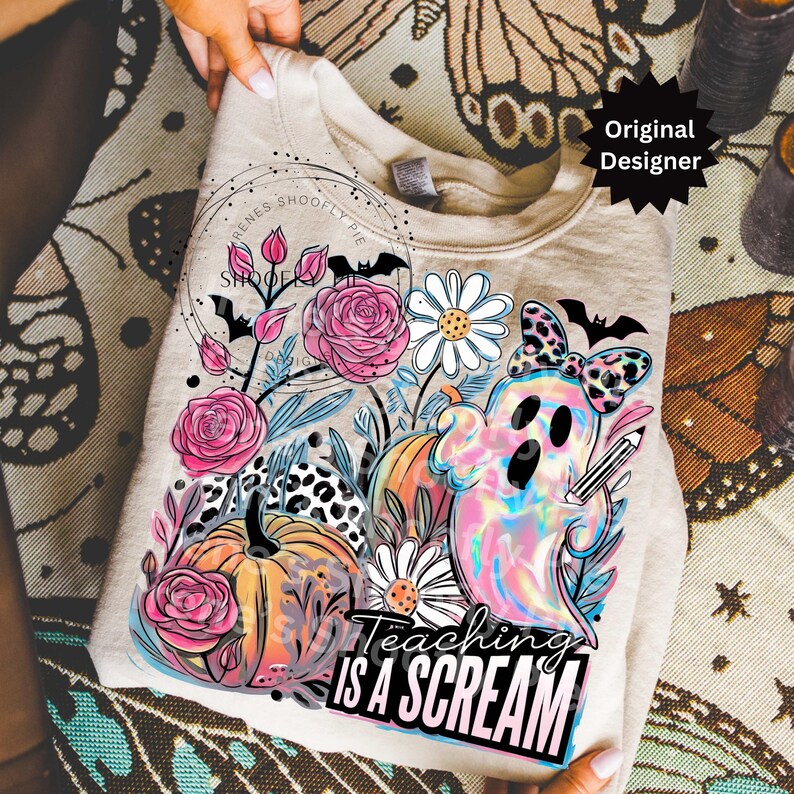 May include: A beige sweatshirt featuring a colorful graphic with a ghost, pumpkins, flowers, and the text "Teaching is a Scream." The design includes pink roses, daisies, and a leopard print bow.