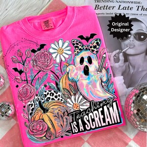 May include: A bright pink Comfort Colors t-shirt featuring a colorful graphic. The design includes a ghost holding a pencil, pumpkins, flowers, and the text "Teaching is a Scream". A bat-shaped bow and other Halloween-themed elements complete the design.