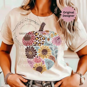 May include: Cream-colored t-shirt featuring a floral acorn design. The acorn is filled with sunflowers, leopard print, and other flowers in shades of pink, yellow, and blue. The text "Original Designer" is visible.