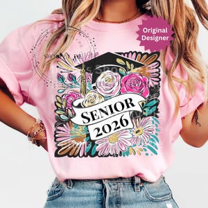 以下が含まれることがあります： 卒業キャップと花柄のデザインが施されたピンクのTシャツ。デザインには、ピンクと白のバラ、デイジー、バナーに「SENIOR 2026」の文字が含まれています。「Original Designer」ステッカーが見えます。