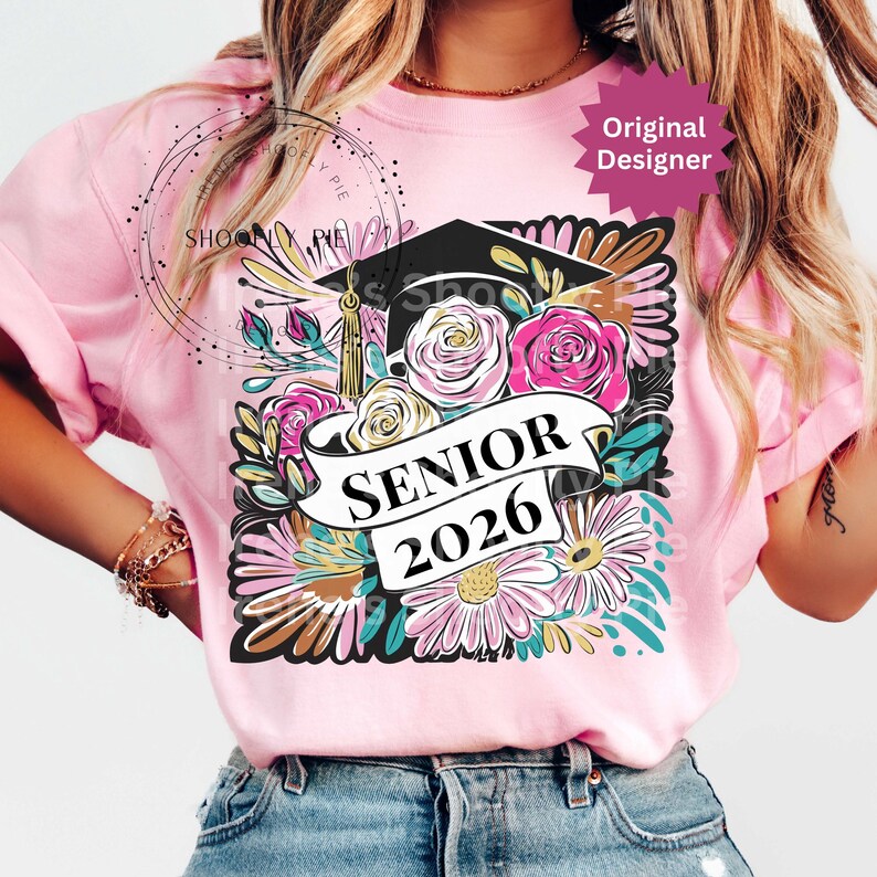 以下が含まれることがあります： 卒業キャップと花柄のデザインが施されたピンクのTシャツ。デザインには、ピンクと白のバラ、デイジー、バナーに「SENIOR 2026」の文字が含まれています。「Original Designer」ステッカーが見えます。