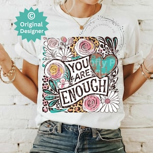May include: White t-shirt with a floral design and the text "You Are Enough" in black lettering. The design also includes a turquoise heart with a distressed look and leopard print.