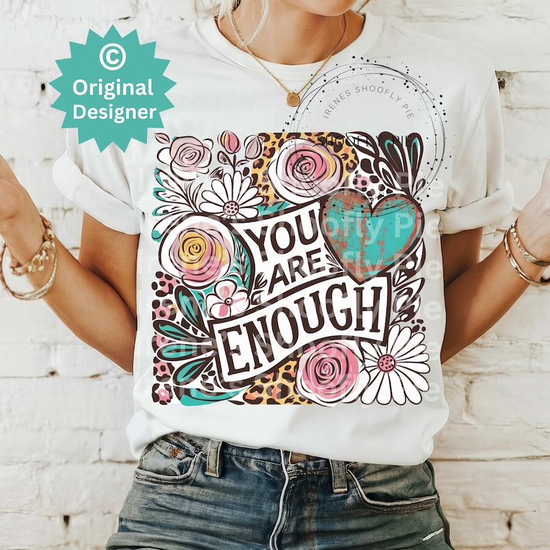 May include: White t-shirt with a floral design and the text "You Are Enough" in black lettering. The design also includes a turquoise heart with a distressed look and leopard print.