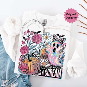 May include: White sweatshirt with a colorful Halloween-themed graphic. The design includes a ghost, pumpkins, flowers, and the text "Teaching is a Scream". The sweatshirt has long sleeves and a crew neck.