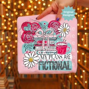May include: Pink t-shirt with a graphic design featuring books, roses, daisies, and a coffee cup. The text "MY PLANS ARE FICTIONAL" is displayed in a teal font. The design includes a bow and the words "Original Designer".