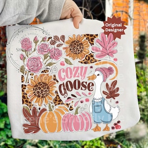 Puede incluir: Un bolso tote blanco con un diseño otoñal. El diseño incluye girasoles, calabazas y un ganso con un peto y un sombrero rosa. También se muestran las palabras "Cozy Goose". El bolso lo sostiene una persona.