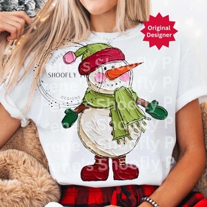 Op de afbeelding: Wit T-shirt met een sneeuwmanontwerp. De sneeuwman heeft een wortelneus, een rode hoed, groene sjaal en wanten en rode laarzen. De tekst "IRENES SHOOFLY DESIGNS" staat op het shirt. Een rode ster met "Original Designer" staat rechtsboven.