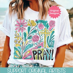 May include: White t-shirt with a colorful floral design, a yellow cross, and the word "PRAY!" in black. The design includes pink, blue, yellow, and green elements. The shirt has rolled-up sleeves.